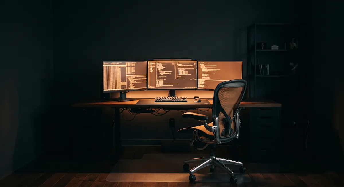 Birou întunecat noaptea, scaun gol și trei monitoare care rulează singure procese automate, lumină portocalie caldă.